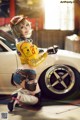 A woman in a yellow jacket and hat posing next to a white car.