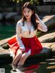 A woman in a red and white dress sitting on a rock by the water.